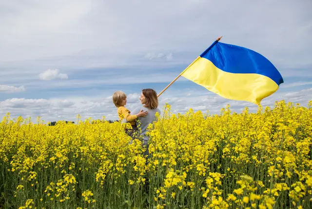 A field of yellow flowers with a woman holding a child and a Ukrainian flag billowing in the breeze