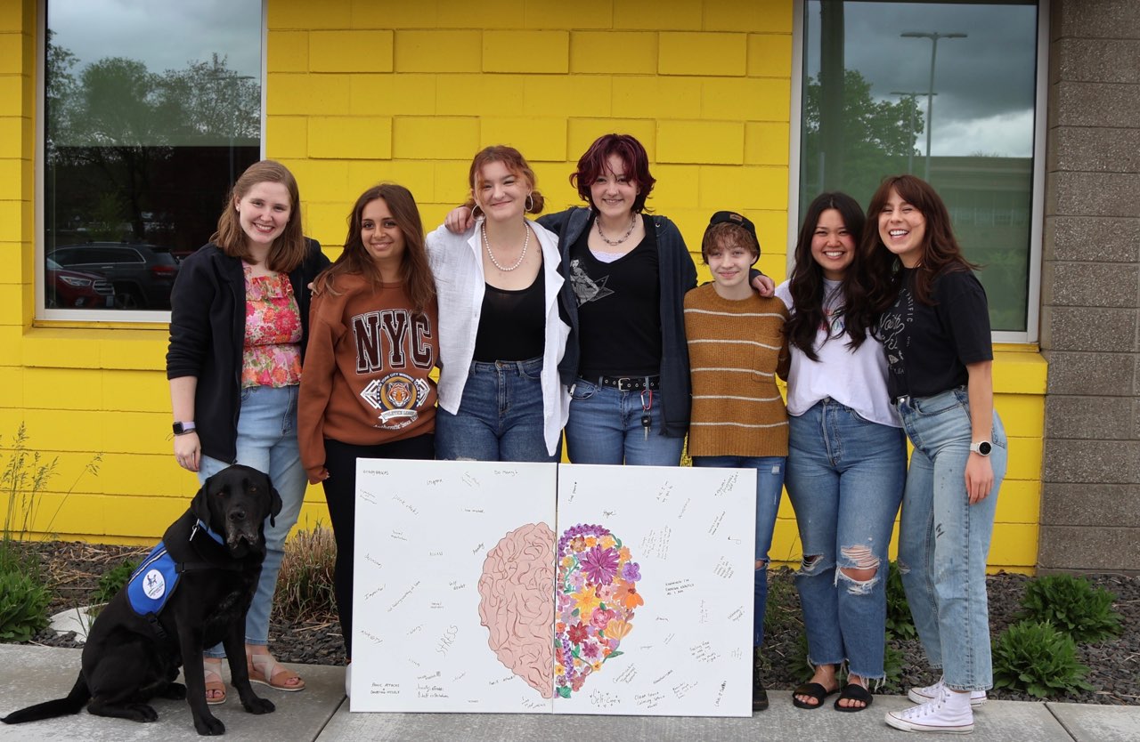 A group of youth and a facility dog are standing together with a poster creation.