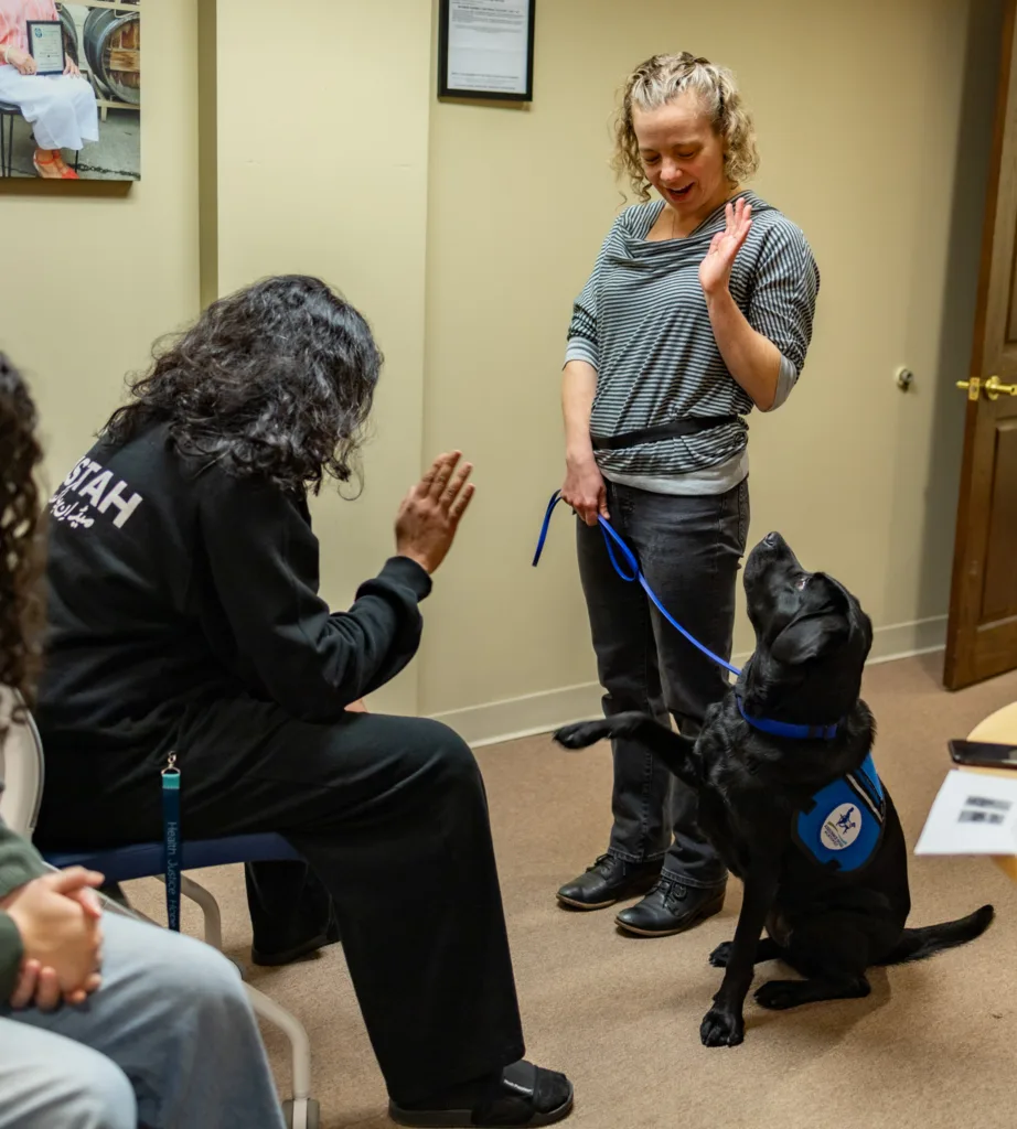 A handler is demonstrating a command with a black service dog who is raising a paw. A person is sitting in front of the two, watching.