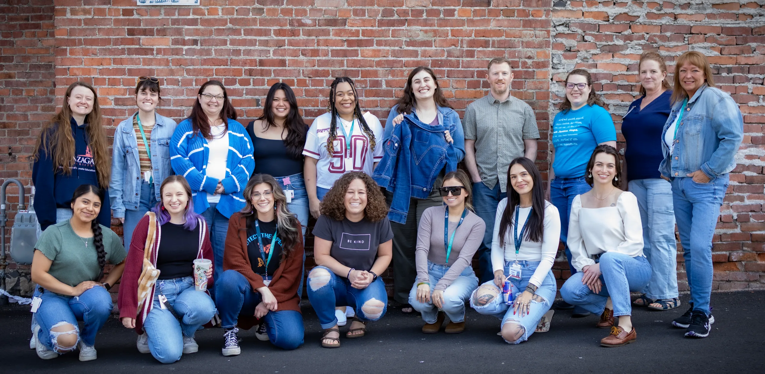 17 people grouped together in front of a brick wall, wearing or showing pieces of denim.